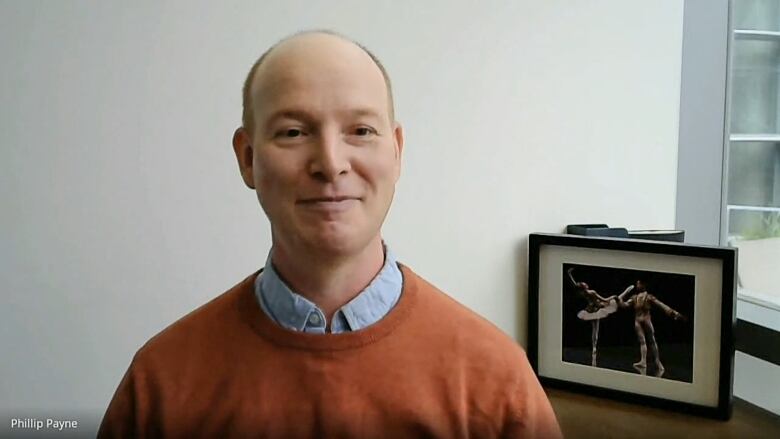 A man in a orange sweater and blue button up smiles at the camera from an office. 