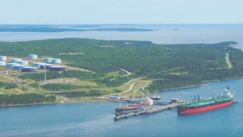 An aerial shot shows an oil and gas storage facility in the distance, with ships on the water in the foreground.