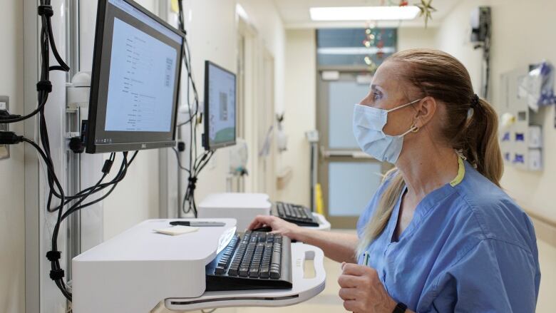 A woman wearing blue hospital scrubs and a medical masks stands in front of a computer monitor in a hospital.