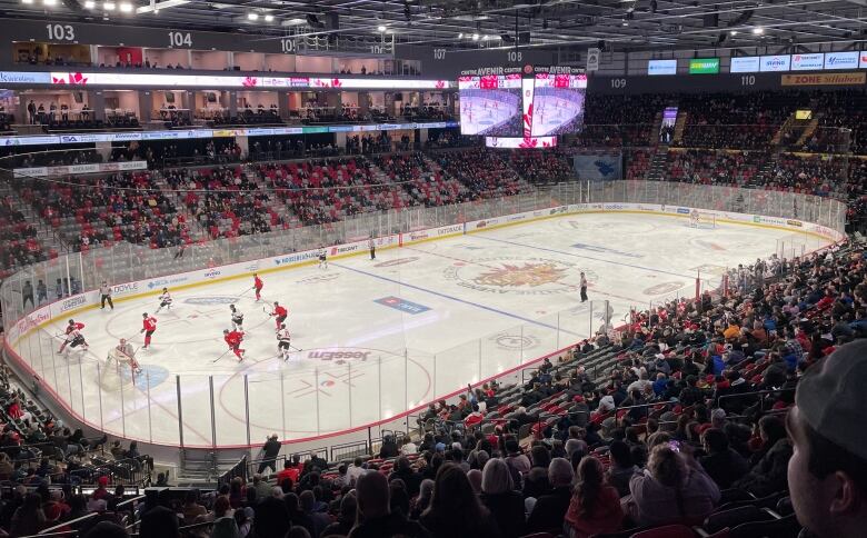 A hockey rink with stands of people surrounding it. Players are concentrated on the left half of the ice.