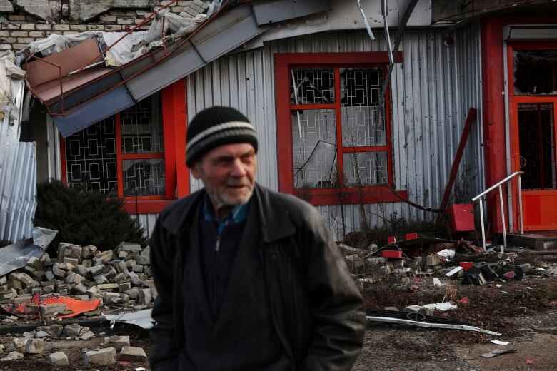 Elderly individual wearing a toque, standing in front of a destroyed home. 