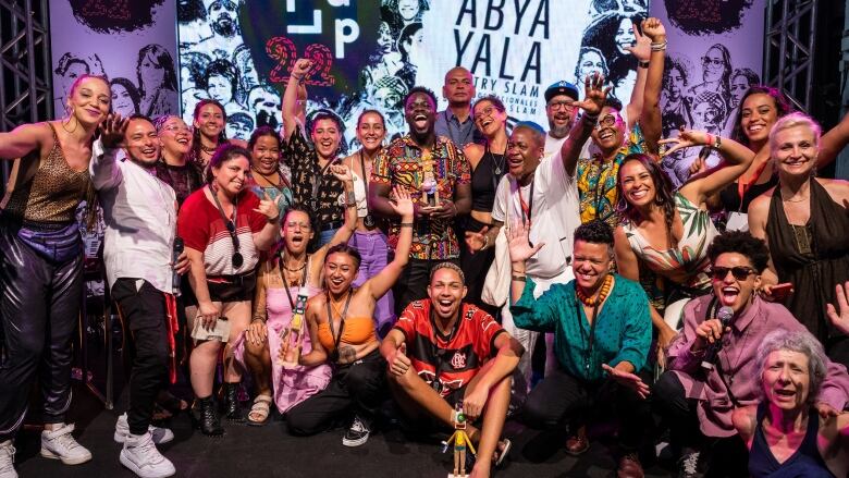 A man holding an award grins while posing with 22 other people for a photo on stage.