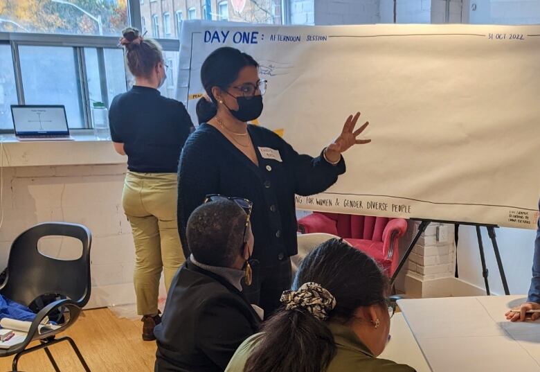 A woman wearing a face mask talks with people sitting at desks. A large sheet of writing paper that says 