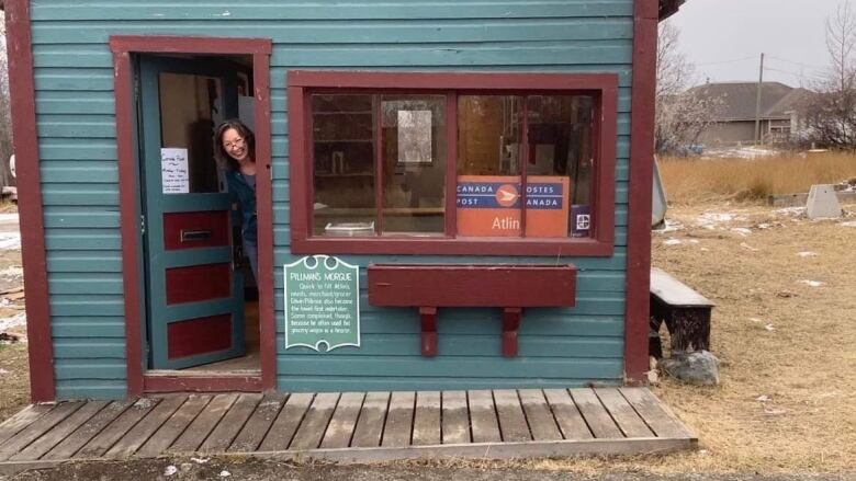 A smiling woman looks out from the open door of a small blue building.