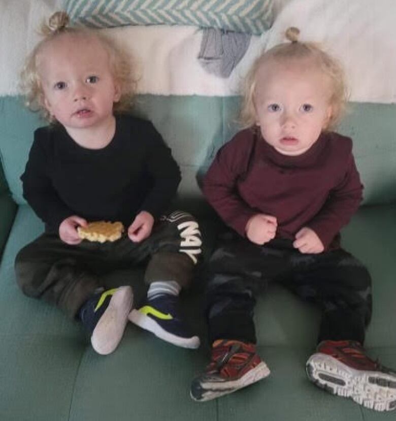 Twin baby boys wearing long-sleeved shirts, pants and sneakers look out while seated on a couch.