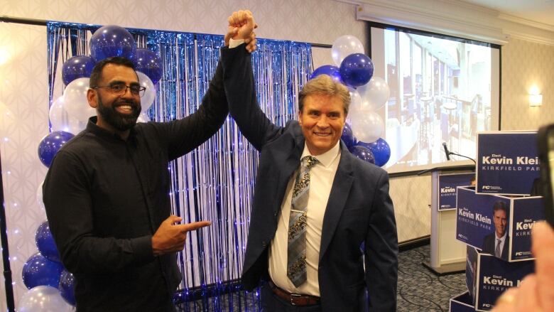 Two men holding each other's arm up with balloons in the background.