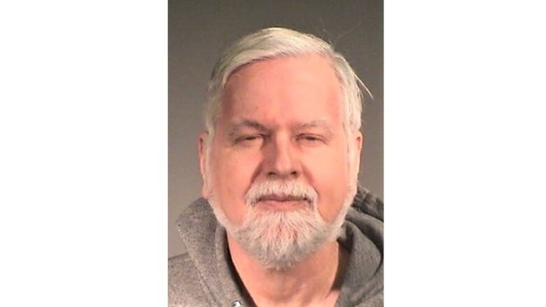 A white man in his 60s with short white hair and white facial hair wearing a grey hooded sweatshirt against a grey background.