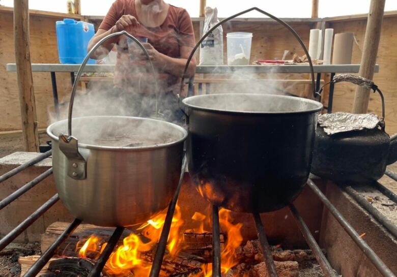 Pots are boiling over a fire as a woman sitting on a picnic bench monitors the fire.