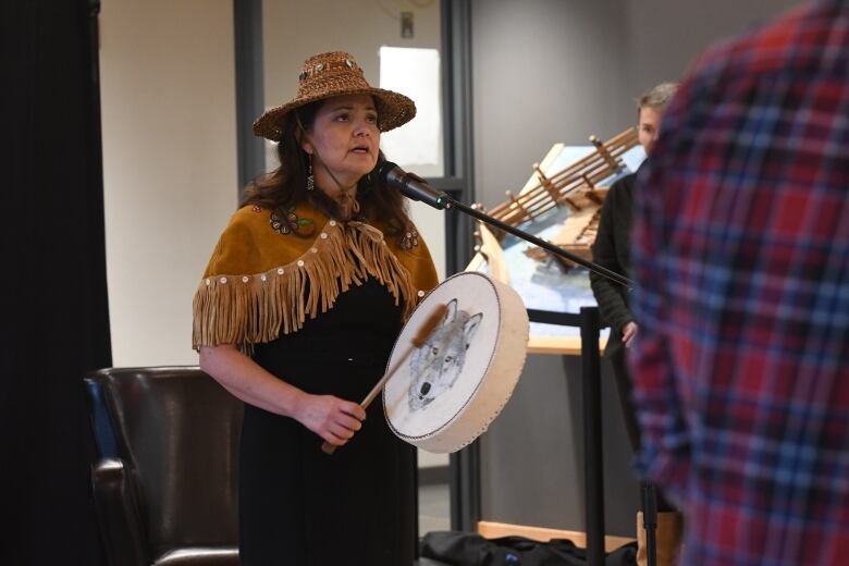 A woman wearing a woven cedar hat and shawl with fringes holds a hide drum decorated with a wolf as she sings into a microphone.