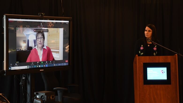 A woman with short hair, glasses and a red shirt is displayed on a TV screen as she appears by Zoom. Another woman with long dark hair stands at a wooden podium next to the TV and is looking at the screen.