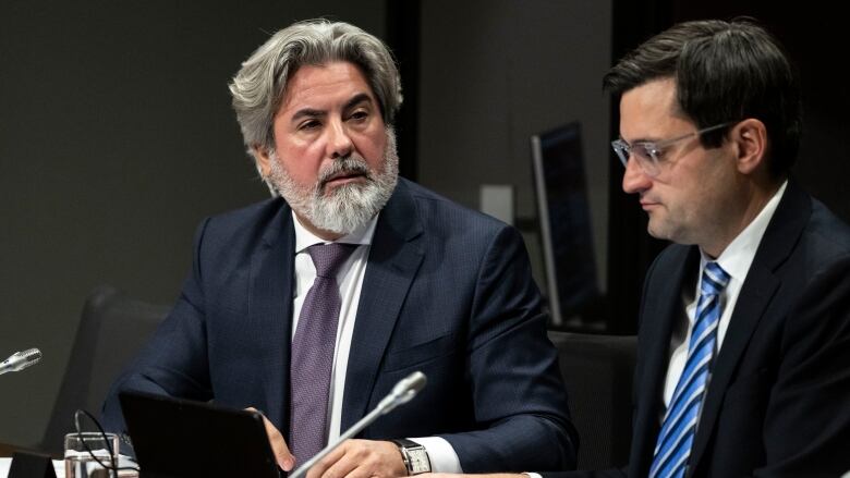 Two men in suits sit alongside each other at a conference table.