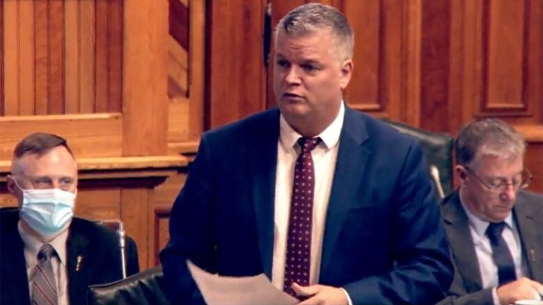 A man standing and speaking in the New Brunswick Legislature, while holding papers.