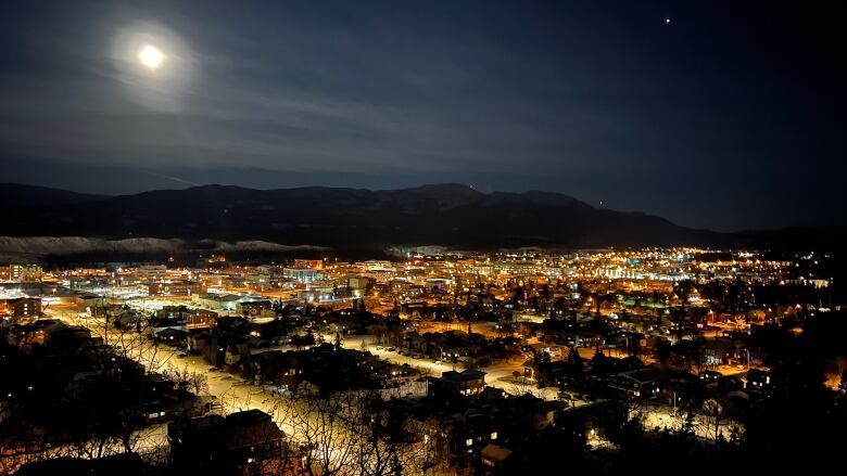 A view of a city at night, with the moon and mountains in the background.