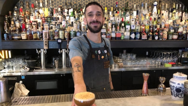 A young man stands behind a beautiful bar with an alcove behind him full of bottles. He's handing a drink toward the camera that looks brown with foam, like and espresso martini. 