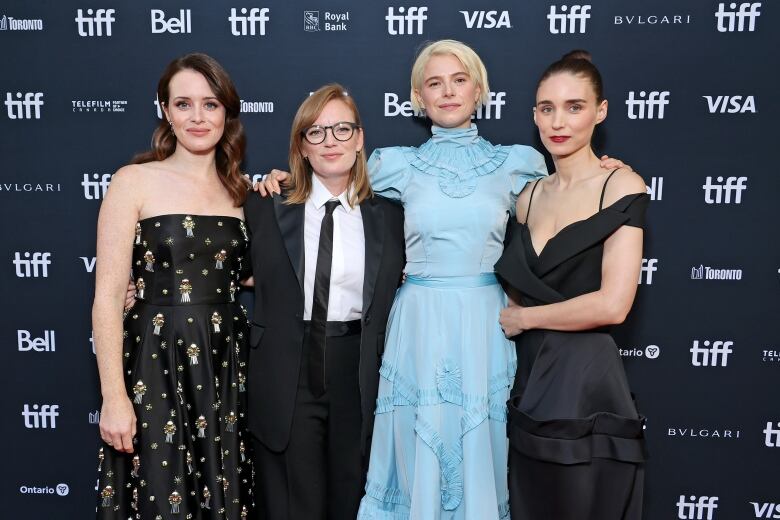 (L-R) Claire Foy, Sarah Polley, Jessie Buckley and Rooney Mara attend the Women Talking premiere during the 2022 Toronto International Film Festival at Princess of Wales Theatre. They are four white women dressed in formal attire and they link arms while posing for the camera against a black-and-white step-and-repeat backdrop branded with logos for TIFF, The City of Toronto, Telefilm Canada and other sponsors.