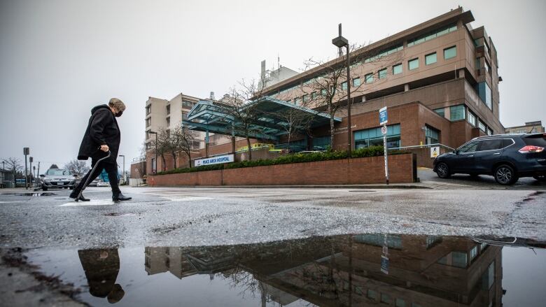 A patient with a cane walks through a crosswalk past a puddle towards Peace Arch Hospital in White Rock, B.C.