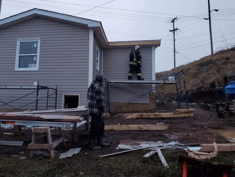 A man is standing on the ground. Another man stands on scaffolding, near a construction site of a new house. 