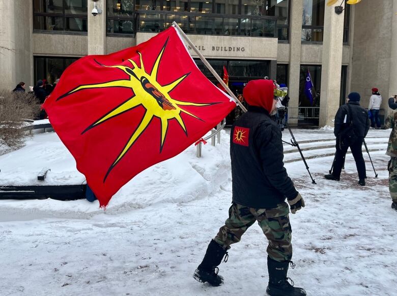 A person wearing black and camouflage clothes carries a warrior flag, which depicts a long-haired Indigenous man over a sunburst atop a red banner, in front of Winnipeg's city hall. The back of his jacket also has a warrior flag on it.