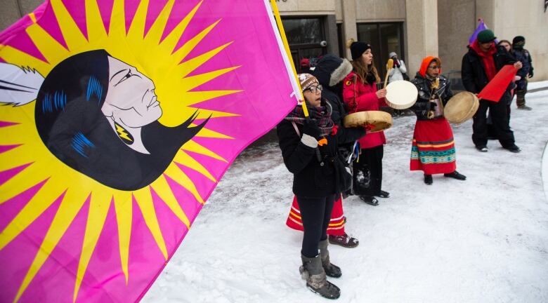 Half a dozen protesters stand on the snow outside Winnipeg city hall on Thursday, three holding drums and one in the foreground holding a large, pink flag with a yellow starburst in the centre, around a drawing of a woman with feather in her long, black hair.