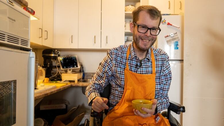 A man in a wheelchair holds up a bowl of soup in a kitchen and smiles.