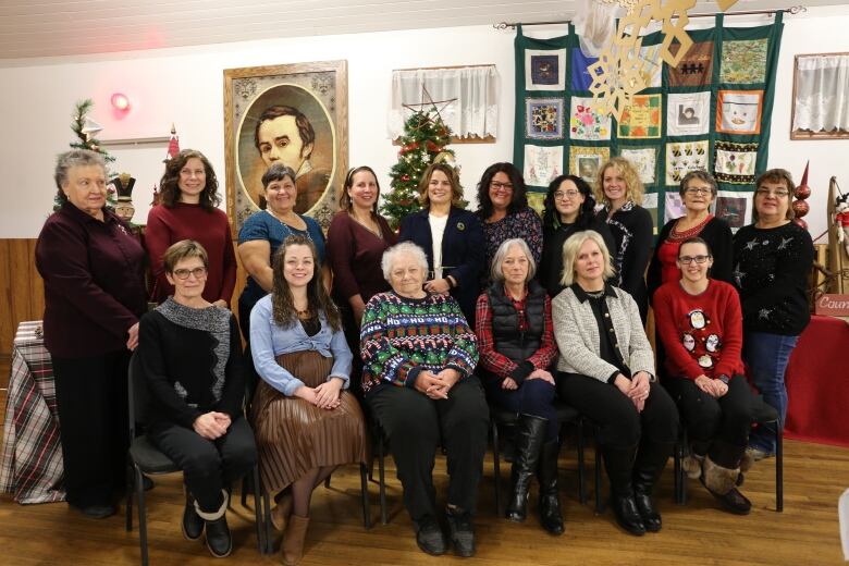 Two rows of women pose for a photo in a hall with Christmas decorations.