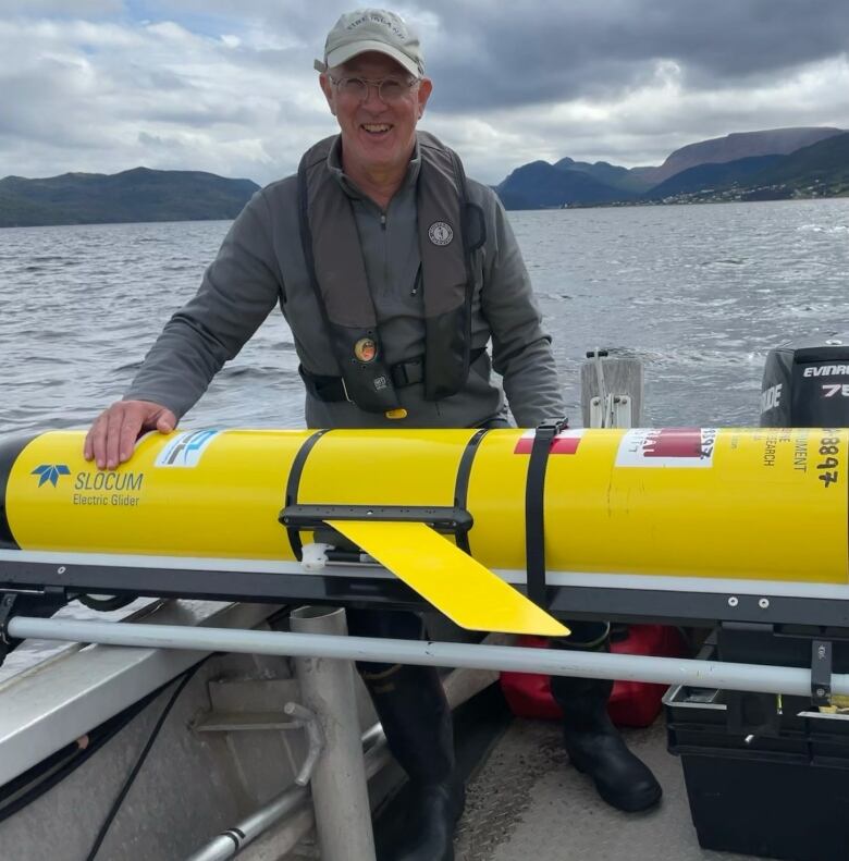 A man in a lifejacket stands in a speedboat while resting his hand on a large yellow underwater drone.