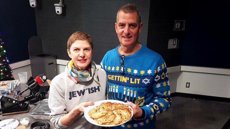 woman and man stand side by side, smiling, holding plate of latkes