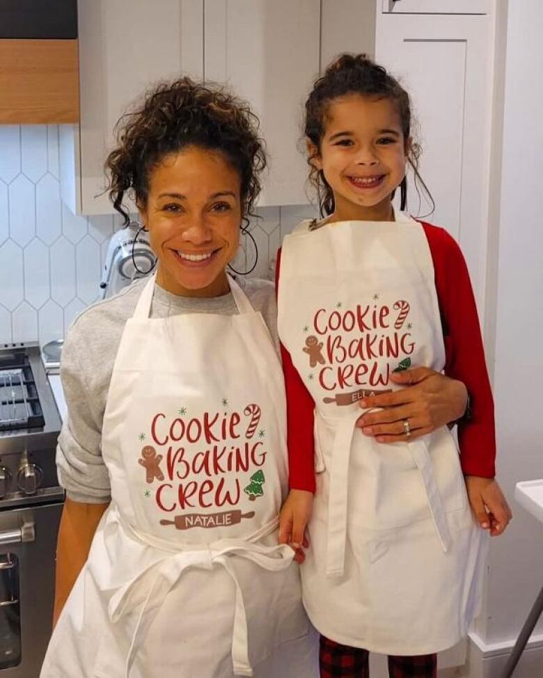 Woman smiling beside female child in a kitchen, both with aprons that read 