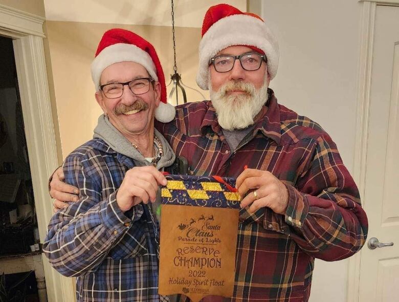 Two men wearing Santa hats hold an award in their hands.