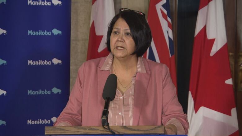 A woman in a pink button-up and blazer and glasses on her head speaks with a pained look on her face in front of a backdrop of Canadian and Manitoban flags.