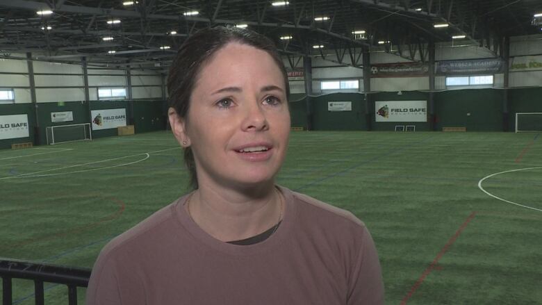 A woman with dark hair and wearing a purple shirt talks before an indoor soccer pitch. 