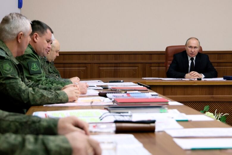 Putin, in a suit and tie, sits behind a table, at a distance from five men in green military uniforms. Their table is perpendicular to Putin's. The table is covered in documents. 