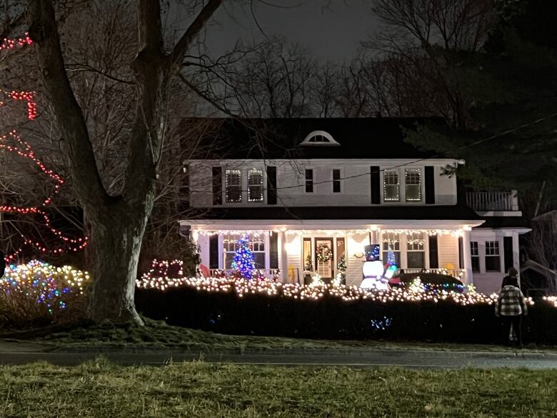 The porch of a white house is illuminated with lights and a Christmas tree and there are white lights on the hedge in front.