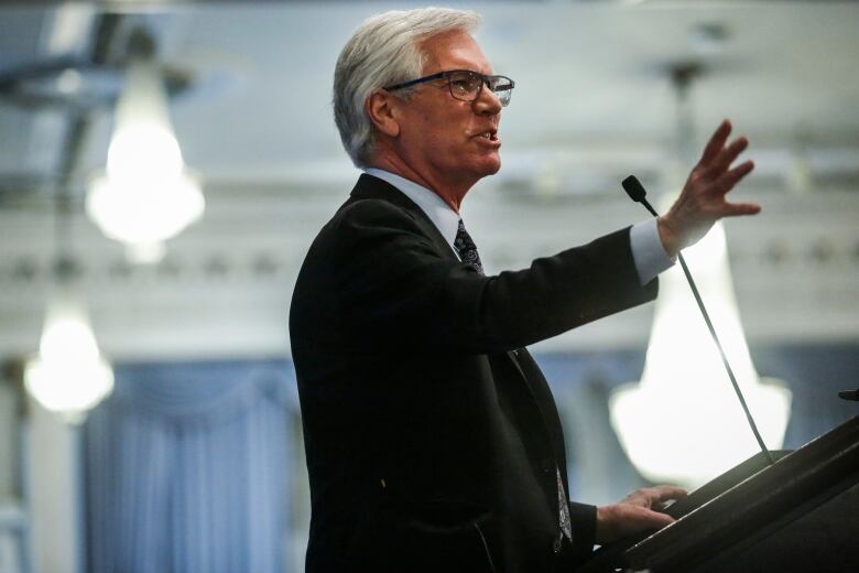 A white-haired man in a suit gestures as he speaks at a podium.