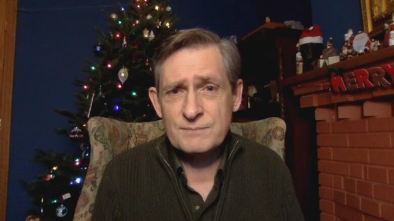 A man looks into the camera. In the background, a Christmas tree and a fire place can be seen.