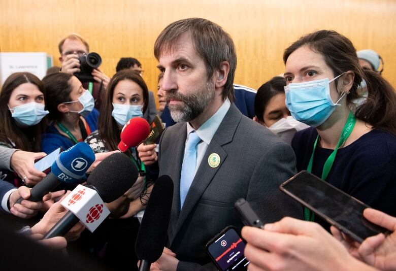 Steven Guilbeault, the minister of Environment, is surrounded by journalists, who are holding up microphones and recorders. Most of the reporters are wearing masks.