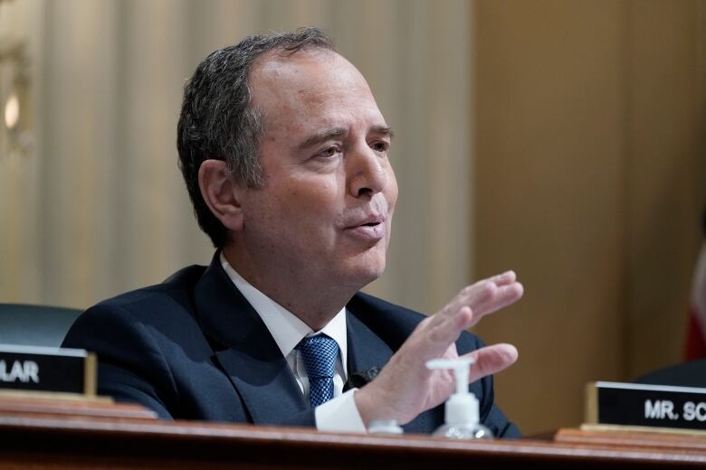 A man is shown speaking while seated behind a table.