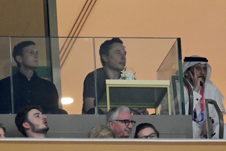 Several men are seen seated inside a stadium. Two of the men are behind a clear partition.