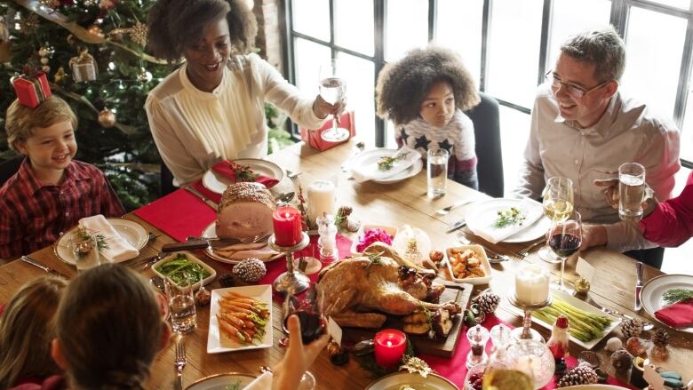 A group of adults and children raise glasses around a holiday table featuring a turkey and side dishes, a Christmas tree in the background.