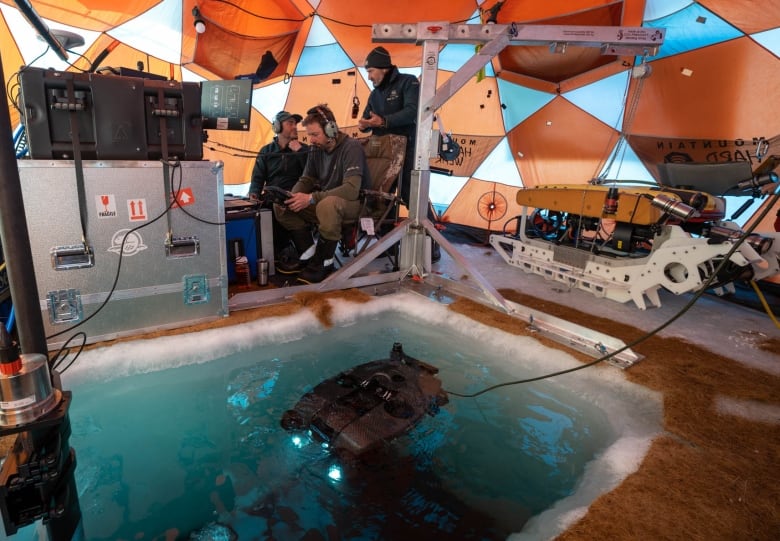 Three men in a domed tent, lots of technical equipment, a square of blue water.