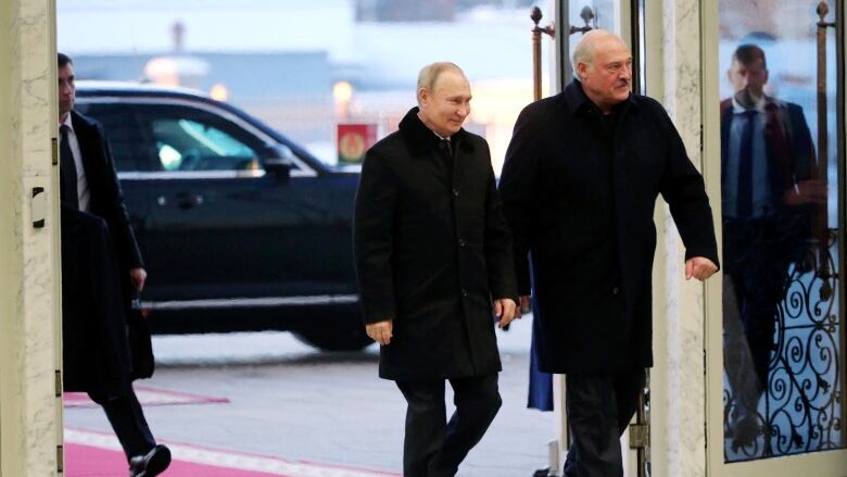 Two men in black coats and black pants walk into a building which appears to have marble floors and walls. The shorter man is smiling. Behind them is a red carpet, and two men who appear to be bodyguards, who are dressed in black suits and black ties.