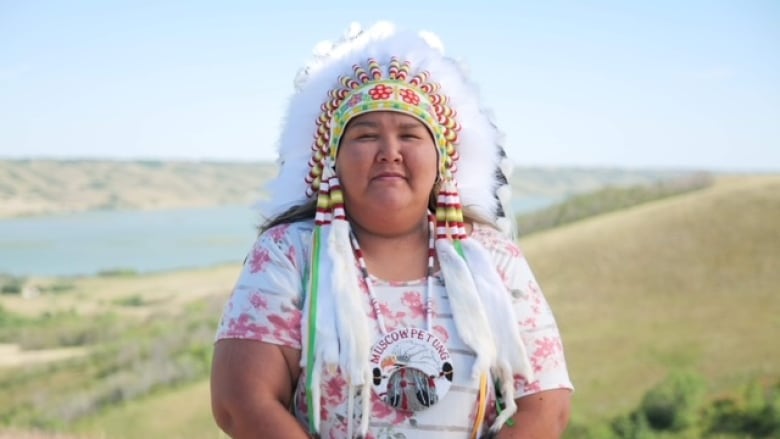 A woman stands in a ceremonial headdress in front of a prairie landscape.