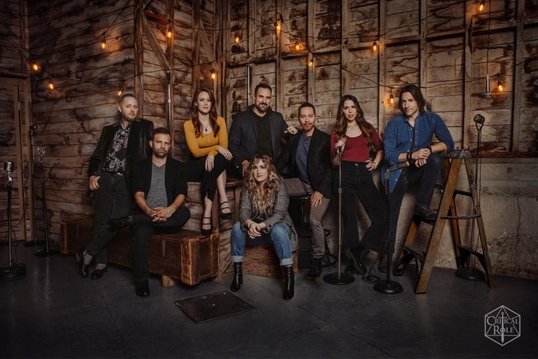 The eight members of the cast of Critical Role pose in front of a wooden backdrop. 