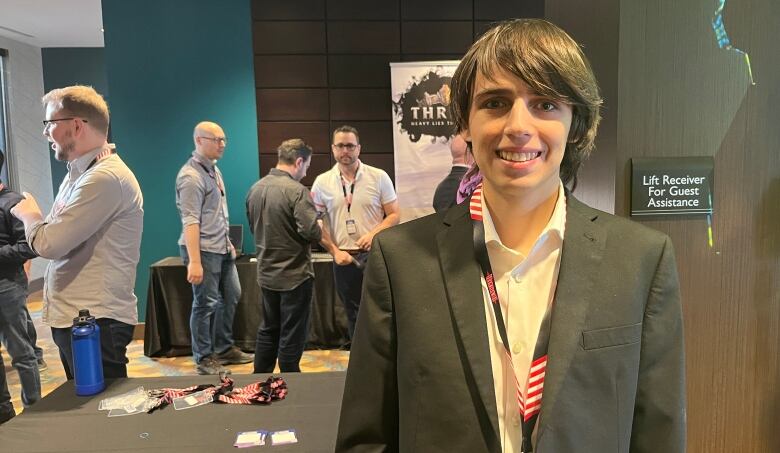 Cole Paskuski stands in a loose off-black blazer and loosened white button-up shirt. He's smiling at the camera , with his ear-lenght brown hair swooped to the side of his face. 