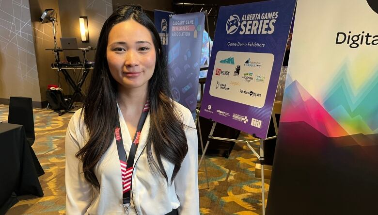 In a white blouse, a woman stands in front of a poster advertising the sponsors for the Alberta Games Series. Her long dark hair twists over her shoulders as the corners of her mouth turn up in a slight smile. 