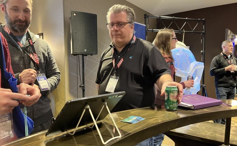 Scott Nye's grey hair is swept back as he watches two men use small controllers to play a video game on a tablet. Nye holds onto a Canada Dry Ginger Ale as he watches the two play. 