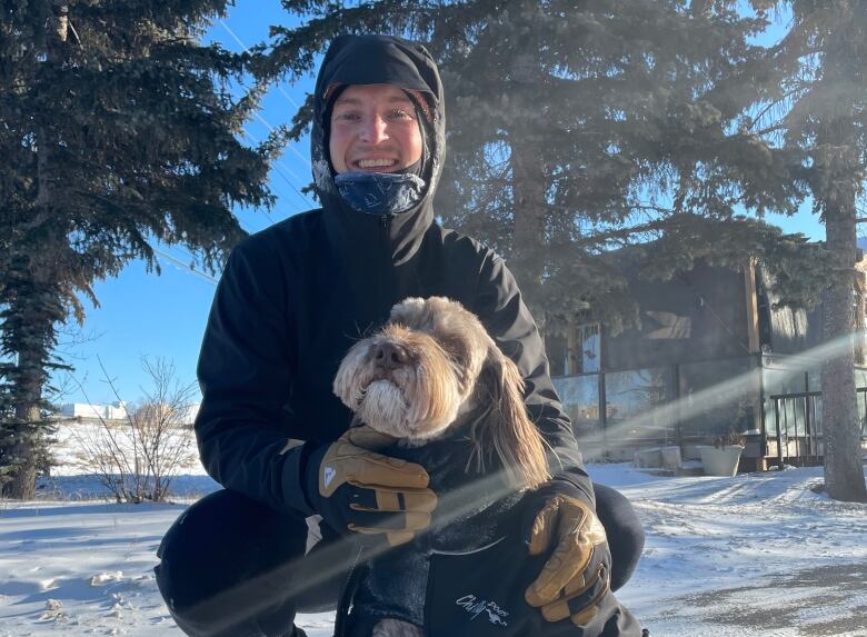 A man wearing a black jacket and dark-coloured running gear kneels next to a sitting dog with fluffy cheecks. 