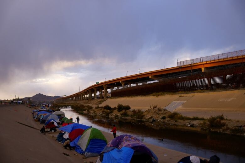 Colourful tents are erected close to the edge of a river. Across the river is a steep hill leading up to an overpass.