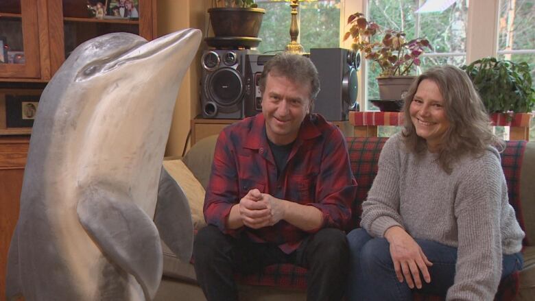 A man in a red shirt sits beside a woman in a grey sweater beside a wooden sculpture of a dolphin. 