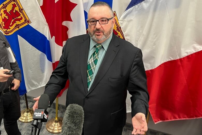 A man with glasses and a beard talks to reporters with outstretched microphones.